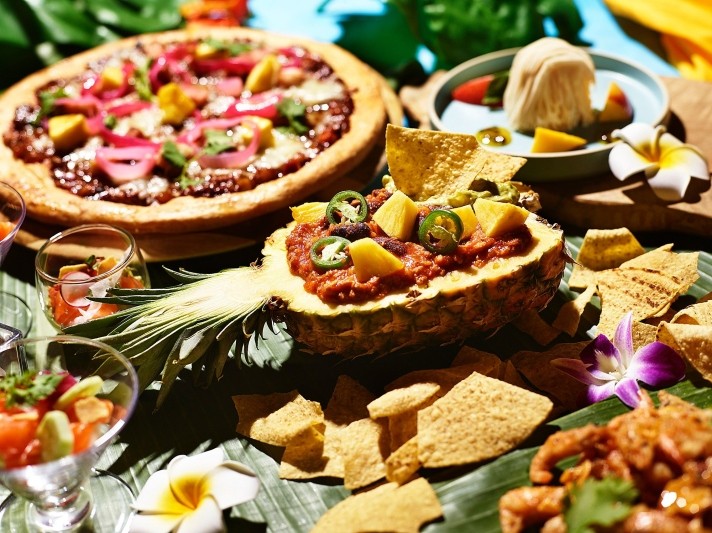 写真：豚肉を乗せたハワイアンプルドポークピザや、トロピカルモンブランなどの夏にピッタリなハワイアン料理が並んでいる様子