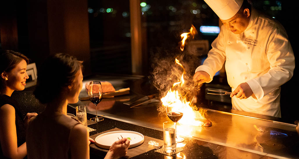 写真：炎を上げて鉄板焼でシェフが調理する様子と女性客二人
